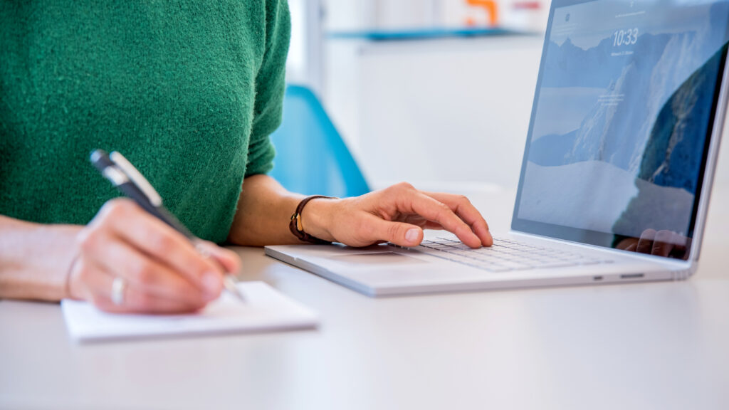 Weibliche Hände. Linke Hand auf Laptoptastatur, Rechte Hand schreibt mit Kugelschreiber.
